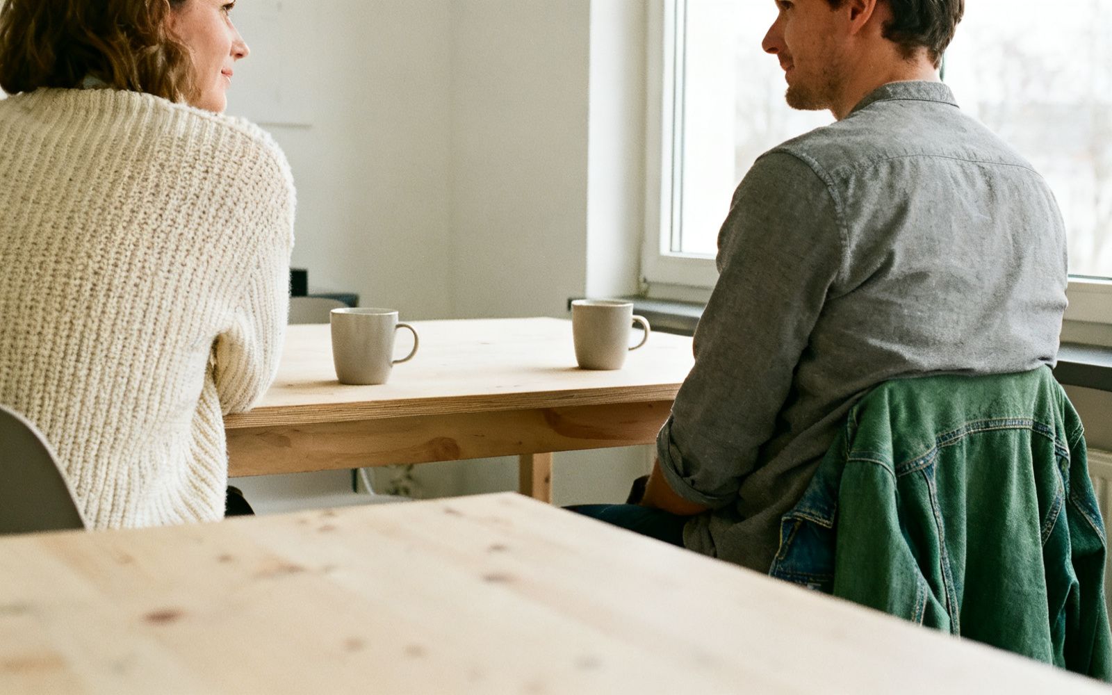 Zwei Menschen sitzen sich an einem Tisch am Fenster gegenüber und sprechen miteinander