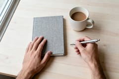 Person sitzt mit einem Laptop an einem hellen Küchentisch, daneben eine Kaffeetasse