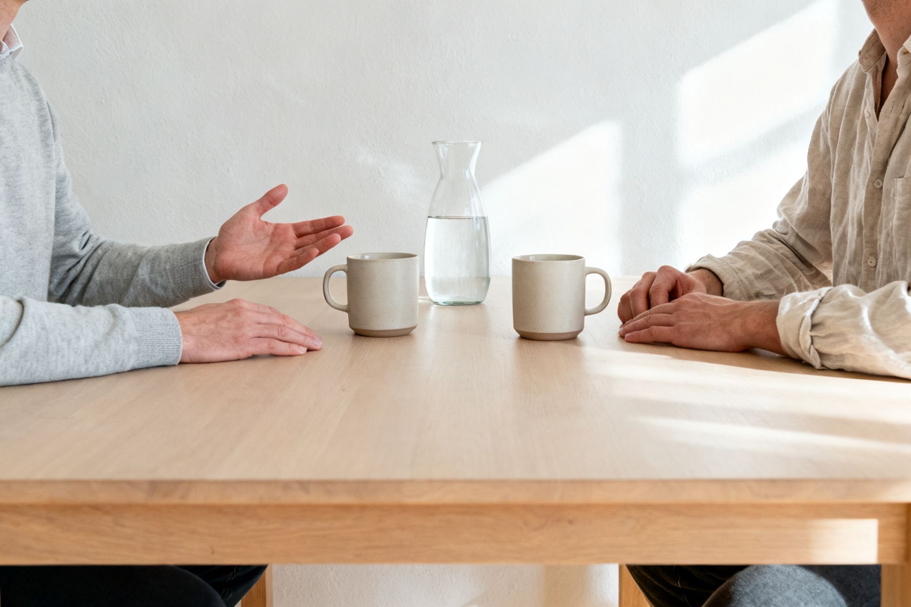 Zwei Menschen sitzen an einem hellen Tisch, zwischen ihnen zwei Tassen, im Hintergrund ein Fenster